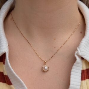 Gold delicate chain necklace with a small flower-shaped pendant featuring a central pearl surrounded by tiny stones, worn on a white-collared shirt.