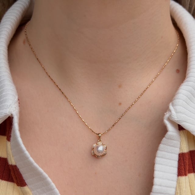 Gold delicate chain necklace with a small flower-shaped pendant featuring a central pearl surrounded by tiny stones, worn on a white-collared shirt.
