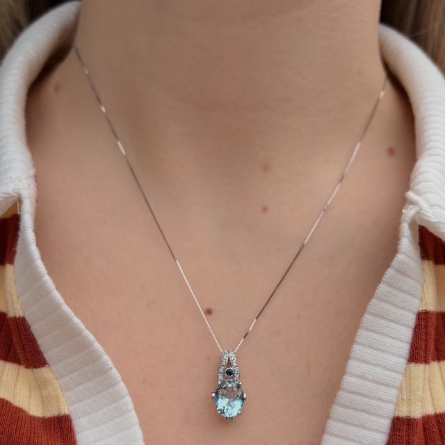 Silver necklace with a blue teardrop pendant on a person’s chest, with a striped shirt visible in the background.
