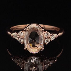 Rose gold ring with a large oval center gemstone and pavé diamonds on the shoulders, on a black reflective surface