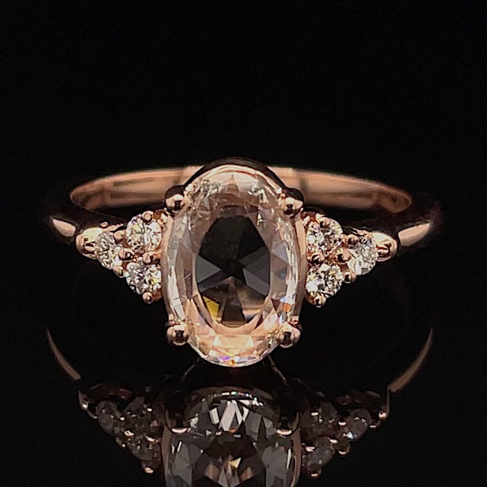 Rose gold ring with a large oval center gemstone and pavé diamonds on the shoulders, on a black reflective surface