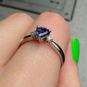 Pear-shaped purple gemstone ring on a finger, flanked by two small diamonds, with a dark metal band and bright green nail nearby.