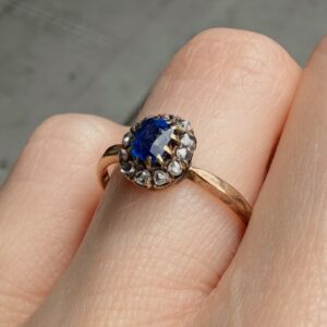 Rose gold ring with a deep blue sapphire encircled by clear diamonds on a finger in close-up.