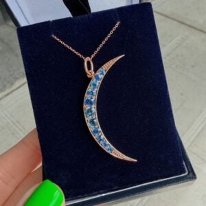 Rose-gold crescent moon pendant encrusted with blue gemstones on a navy velvet display, hanging from a delicate chain.