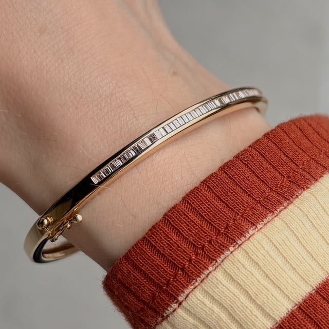 Gold bangle bracelet with a row of baguette-cut stones on a wrist wearing a red-and-cream striped sleeve.