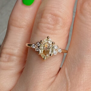 Gold engagement ring with a large oval pale yellow center diamond surrounded by smaller round diamonds, worn on the finger.