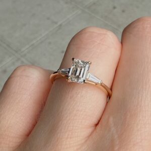 Gold ring with a rectangular emerald-cut diamond centerpiece and baguette side stones, worn on a finger.