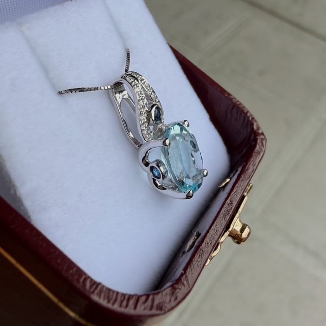 Silver necklace with an oval aqua-blue gemstone pendant surrounded by small diamonds, resting in a burgundy jewelry box.