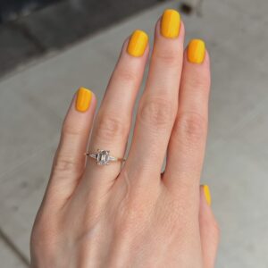 Hand with bright yellow manicured nails; a silver ring with a central rectangular diamond and small side stones on the ring finger, outdoors on concrete pavement