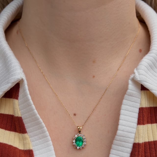Gold chain necklace with an oval emerald pendant surrounded by diamonds, resting on a striped collared shirt.