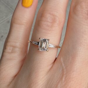 Rose gold ring with a rectangular emerald-cut diamond flanked by baguette diamonds on a finger.