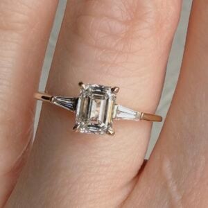 Engagement ring with emerald-cut diamond center and tapered baguette side stones on a slim gold band.