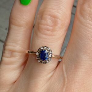 Gold ring with oval blue gemstone surrounded by small diamonds on a finger with green-painted nail polish.