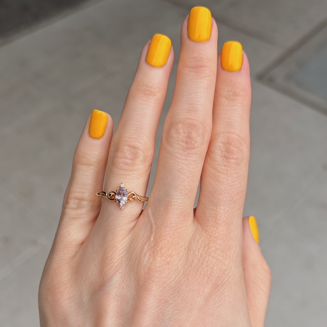 Close-up of a hand with bright yellow-painted nails wearing a gold ring with a pink marquise-cut gemstone on the ring finger.