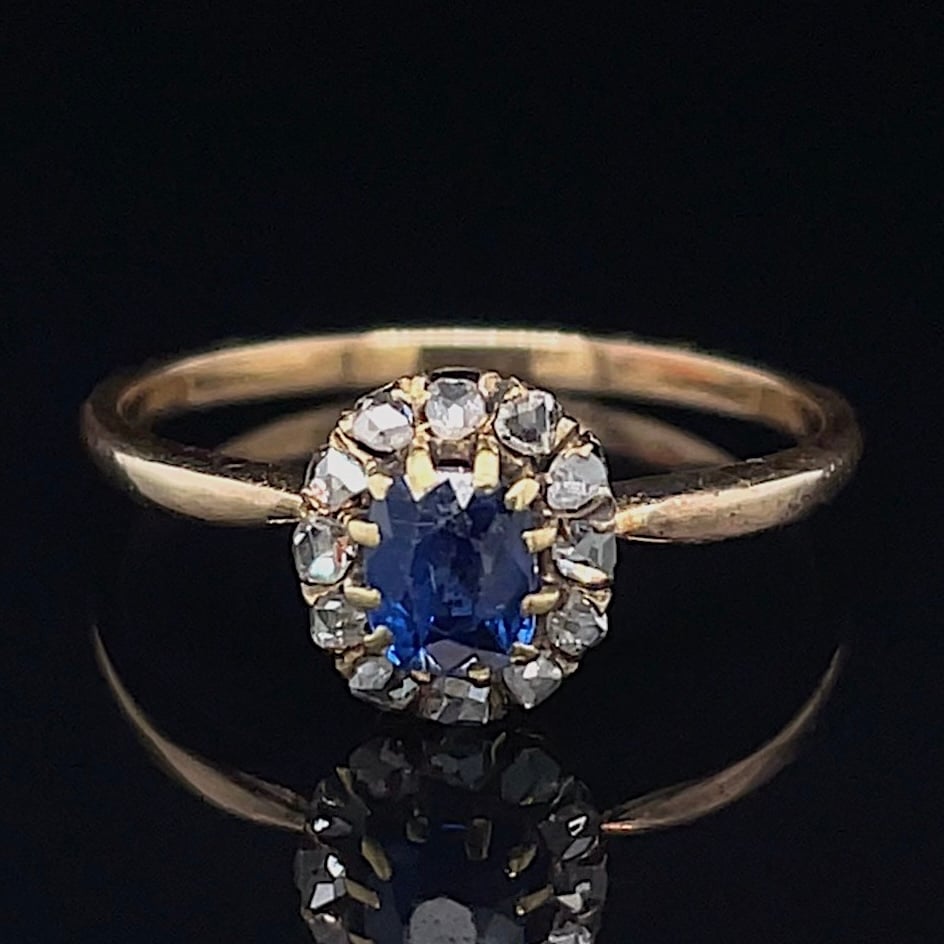 Gold ring with an oval blue sapphire haloed by small diamonds on a yellow-gold band, set against a dark background.