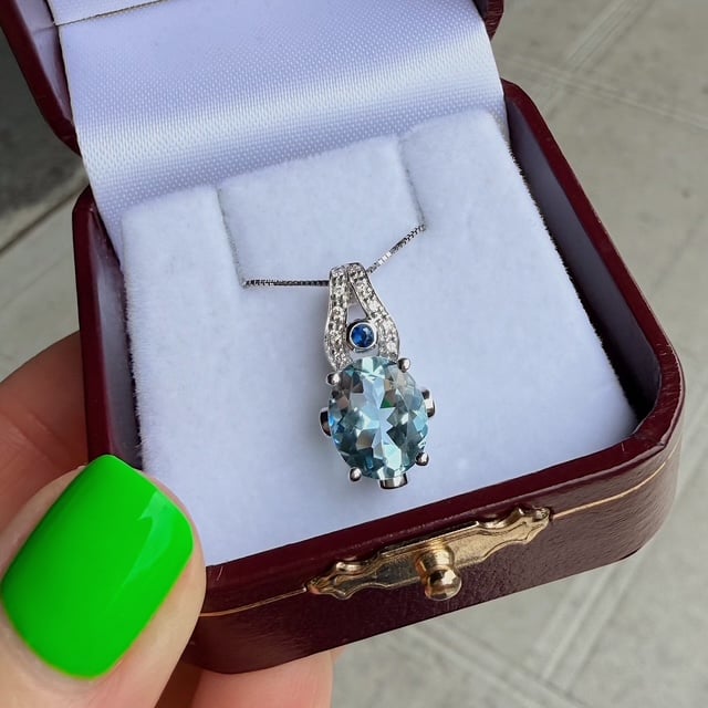 Pendant necklace with an oval light-blue gemstone and diamond accents in a burgundy jewelry box.