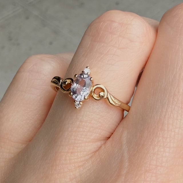 Gold ring with an oval pink gemstone held by prongs, flanked by tiny diamonds and wrapped by decorative scrollwork on a finger.