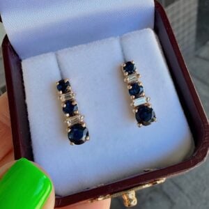 Pair of gold drop earrings featuring deep blue sapphires and clear stones in a white cushioned jewelry box.