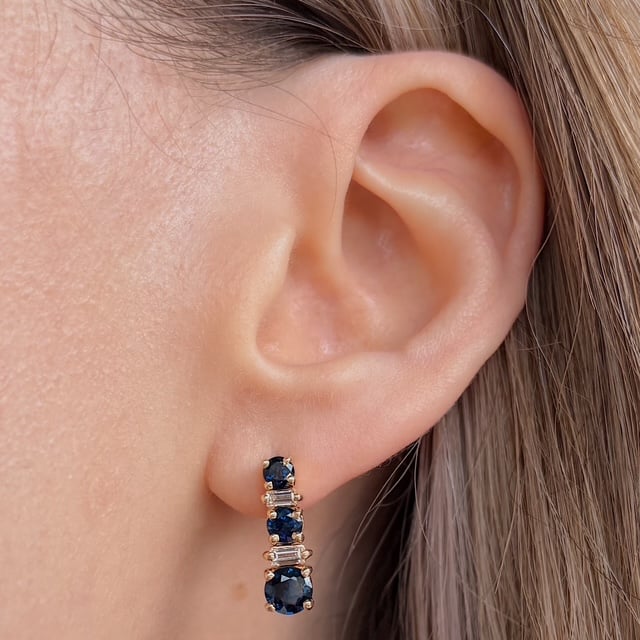 Close-up of the ear with a gold drop earring: alternating dark blue sapphires and clear baguette stones.