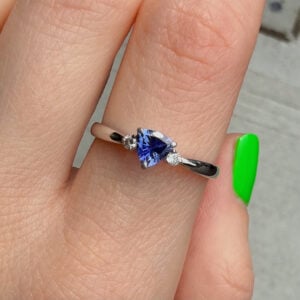 Close-up of a ring on the finger: a blue pear-cut gemstone flanked by two small diamonds on a silver band, with a bright green nail visible nearby.
