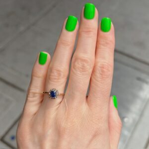 Hand with bright green manicured nails, showcasing a gold ring with an oval blue sapphire surrounded by small diamonds on the ring finger.