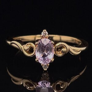 Gold ring with an oval lavender gemstone and decorative scrolling on the band against a black background.