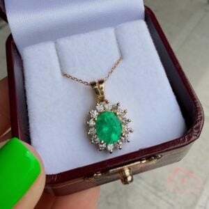 Gold pendant with a large oval emerald surrounded by a diamond halo, resting in a white cushioned jewelry box