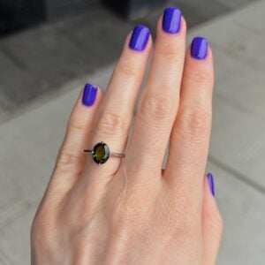 Hand with purple nail polish wearing a silver ring with an oval olive-green gemstone on the ring finger amid a neutral background.