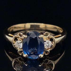 Gold ring with an oval blue sapphire centerpiece flanked by two small diamonds on a decorative gold setting, placed on a black reflective surface.