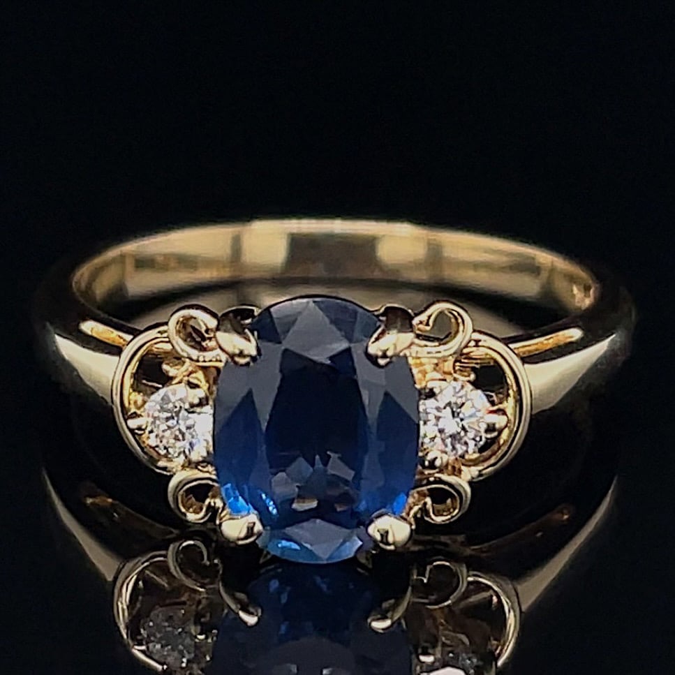 Gold ring with an oval blue sapphire centerpiece flanked by two small diamonds on a decorative gold setting, placed on a black reflective surface.