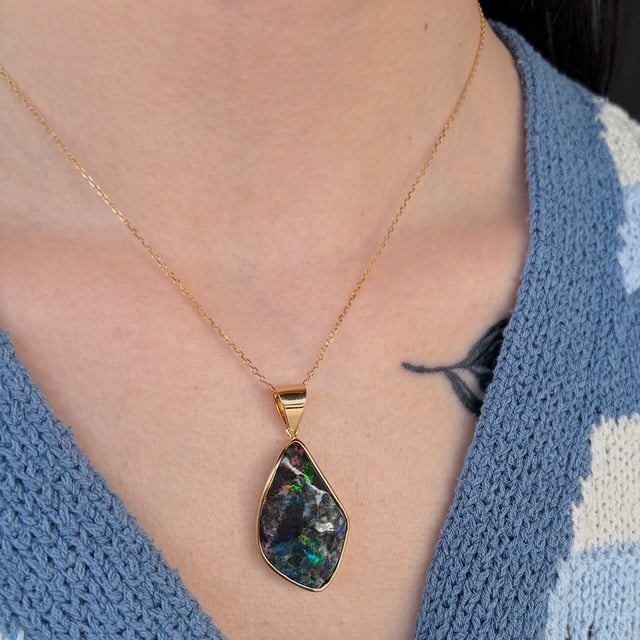 Gold teardrop pendant necklace with a dark opal-like stone on a delicate chain, worn at the neck. A blue knit sweater and a small tattoo are visible nearby.