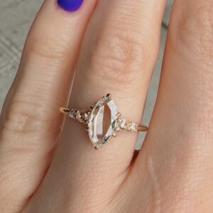 Rose-gold ring with a marquise-cut clear center stone and small side stones, worn on a finger with purple nail polish visible in the background.
