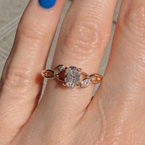 Rose gold ring with a central diamond cluster shaped like a flower, flanked by leaf-shaped pavé diamonds on the band, worn on a finger with blue nail polish.”,“Close-up of a rose-gold ring featuring a diamond floral centerpiece and leaf accents on the band.”,“Jewelry: central diamond cluster on a rose-gold ring with leaf settings, on a finger.”