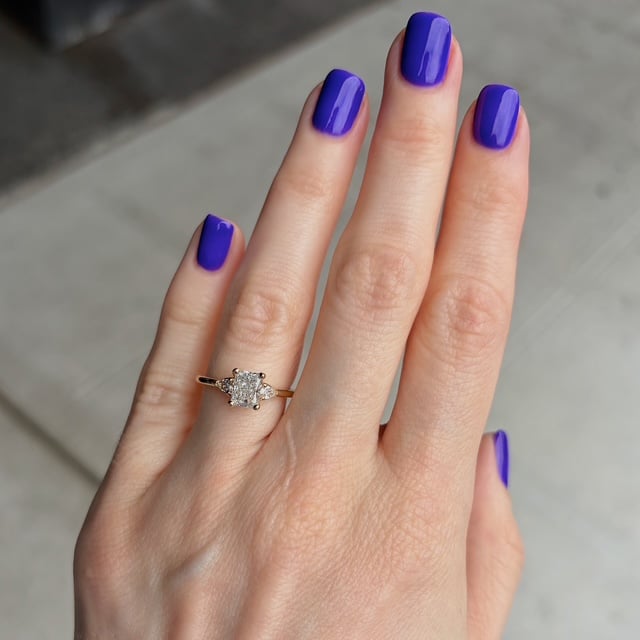Close-up of a left hand with glossy purple nails wearing a diamond engagement ring on the ring finger, featuring a central stone with smaller surrounding stones on a gold band