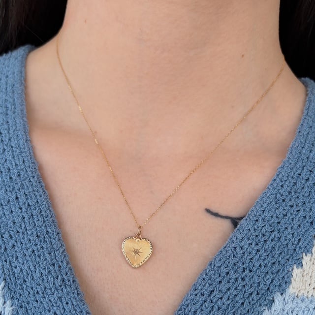 Gold heart pendant on a delicate gold chain resting on a blue knit top; heart features a small starburst design in the center.