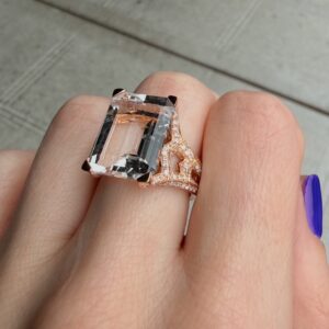 Rose gold ring with a large emerald-cut center stone and diamond-accent band on a finger with purple nail polish.