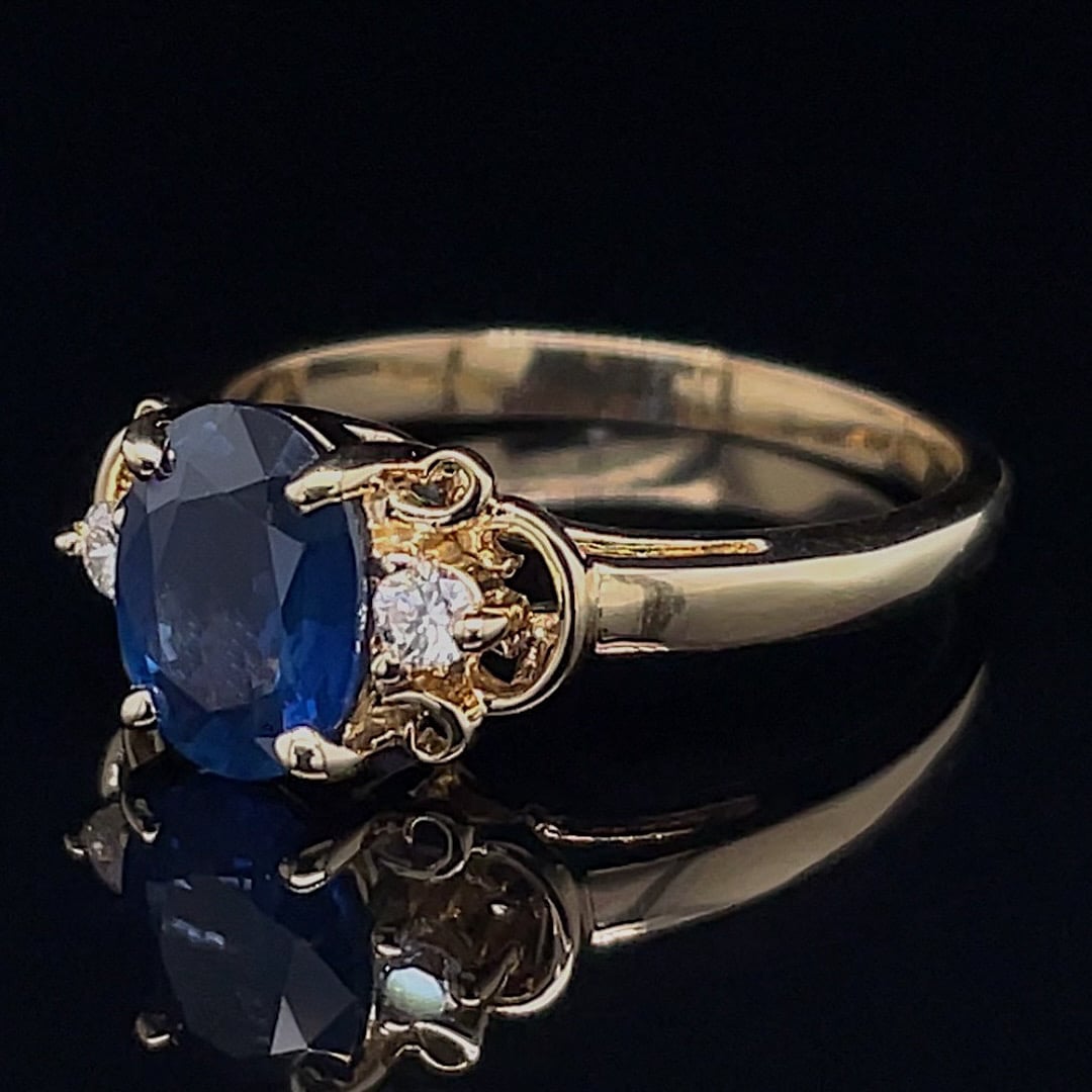 Gold ring with a large oval blue sapphire center, flanked by small diamonds, on a dark reflective surface/double-tone setting