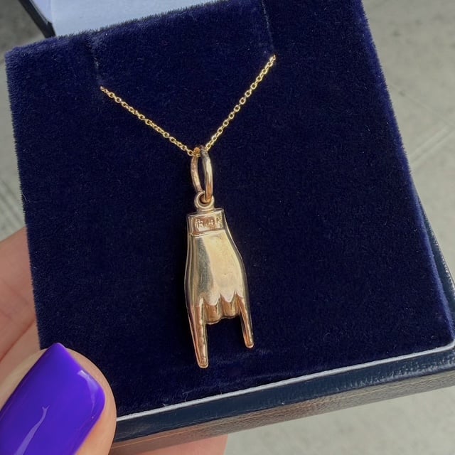 Gold necklace with a pendant shaped like a hand showing the raised middle finger, displayed in a blue velvet jewelry box.
