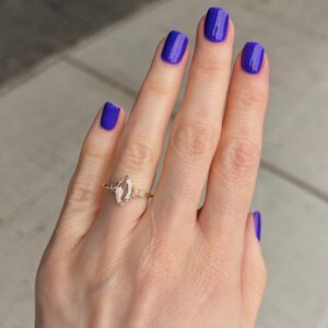 Hand with manicured purple nails wearing a gold ring with a marquise-cut center stone outdoors.