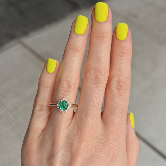 Hand with bright yellow nail polish wearing a ring featuring a green emerald center stone surrounded by small diamonds.