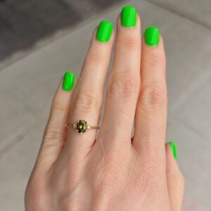 Hand with neon green nail polish wearing a delicate ring with a green gemstone on the ring finger.