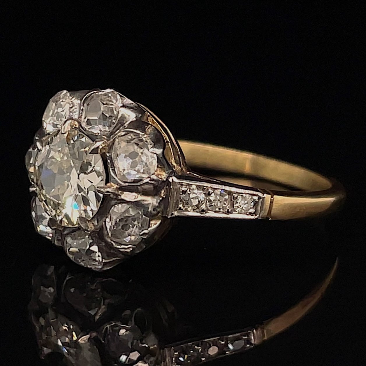 Gold ring featuring a large diamond cluster arranged in a floral-like centerpiece, with pavé diamonds along the band on a black background.