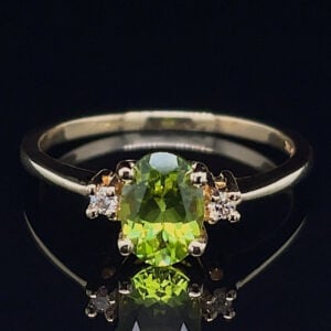 Emerald-green central gemstone ring with two diamond side stones on a metallic band, set against a black reflective background.