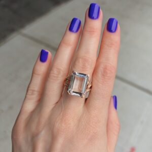 Hand with bright purple nails wearing a large rectangular clear gemstone ring on the ring finger with gold band detailing.