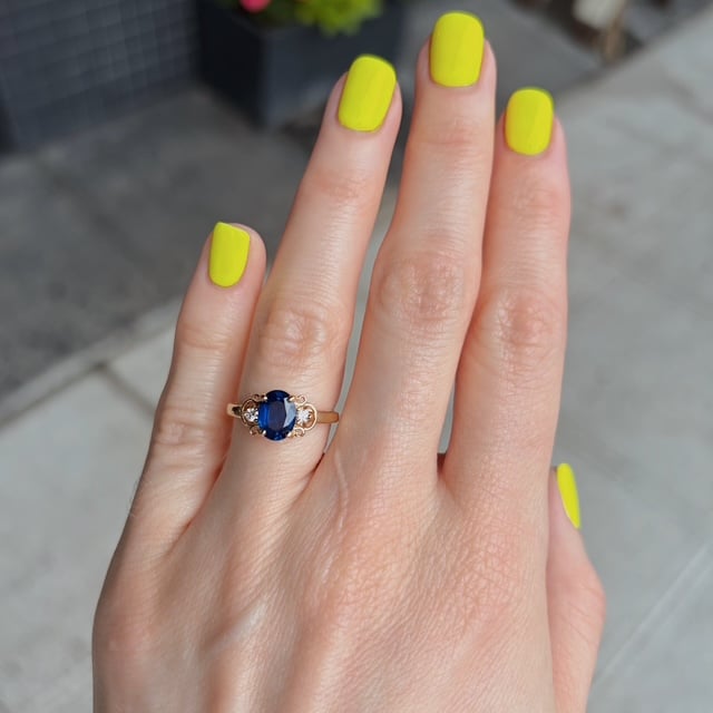 Close-up of a hand with neon yellow nails and a gold ring featuring a blue oval gemstone on the ring finger.