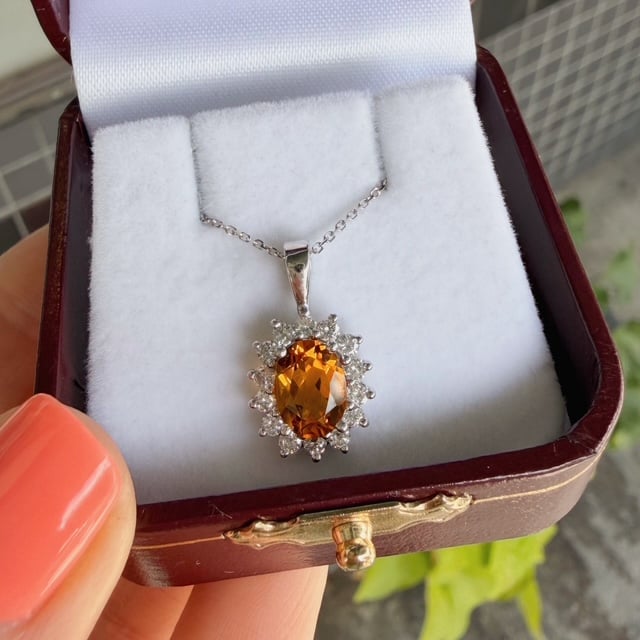 Oval orange gemstone pendant with a diamond halo on a silver chain, in a white cushioned jewelry box.