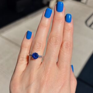 Ring with a round blue sapphire center stone flanked by baguette diamonds on a silver band, worn on a hand with bright blue nail polish.