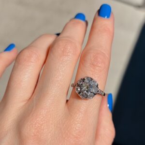 Rose gold halo engagement ring with a central diamond surrounded by smaller diamonds on a finger with bright blue nails and a pale background.