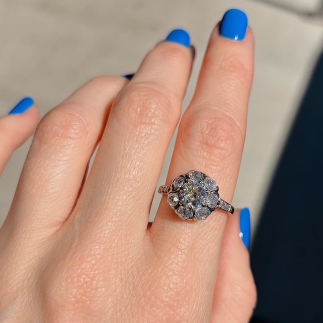 Rose gold halo engagement ring with a central diamond surrounded by smaller diamonds on a finger with bright blue nails and a pale background.