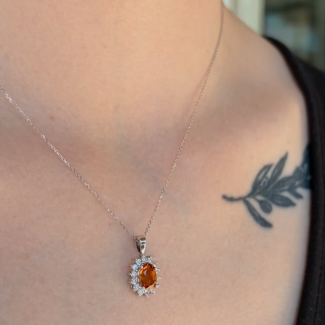 Silver chain necklace with an orange oval gemstone pendant surrounded by diamonds, resting on the chest near a leafy tattoo.